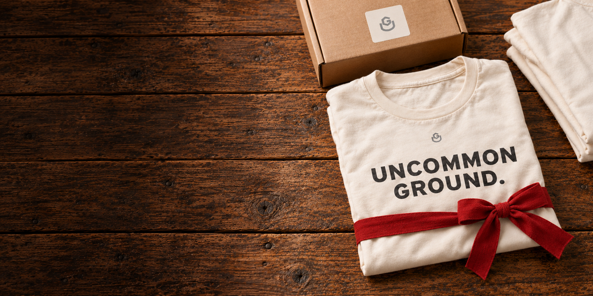 Folded Uncommon Ground tee tied with a red ribbon beside a UG gift box on rustic wood